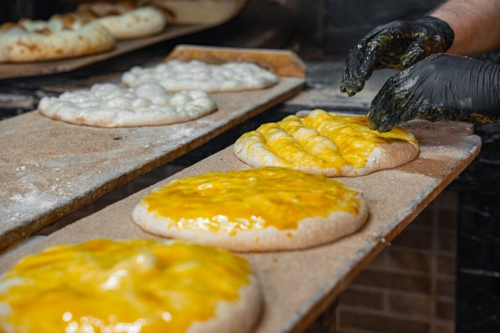 A baker's hand in gloves shaping cheese-covered flatbreads on a wooden board ready for the oven.