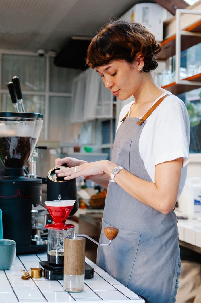 Pensive woman barista with short brown hair in gray apron preparing coffee through paper filter in cafe